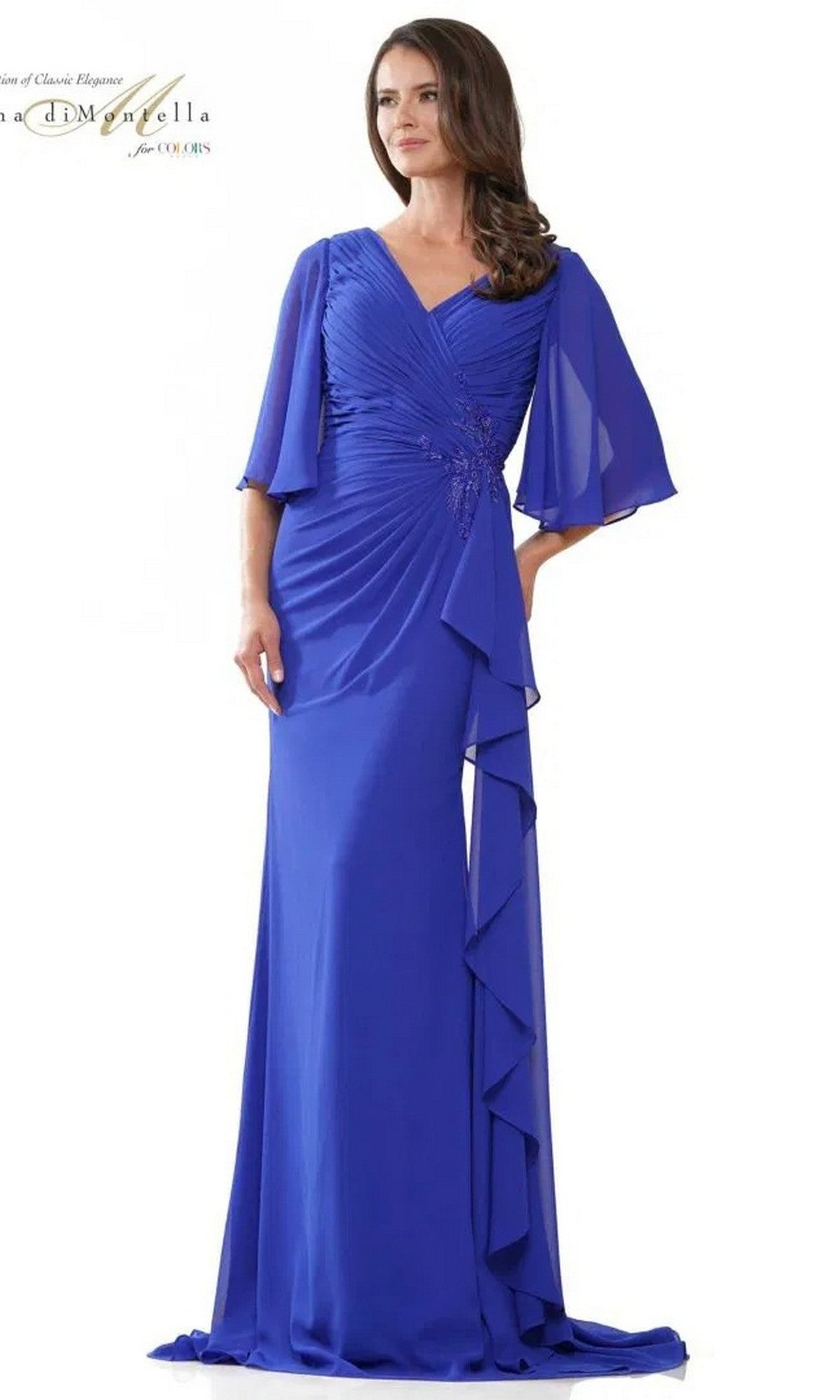 A woman models the Rina Di Montella RD2935 long formal dress in sapphire, with a draped, ruched bodice and flowing sleeves, standing against a plain white background.